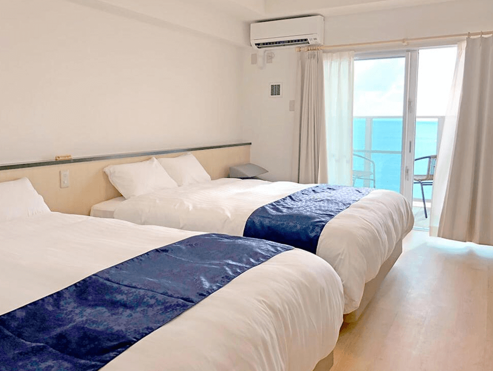 【名護住宿】Hotel Sakurano Familia Nago 海景雙床房，房間簡約明亮，設有兩張雙人床與藍色床尾巾，落地窗外可直通陽台，欣賞碧藍海景。