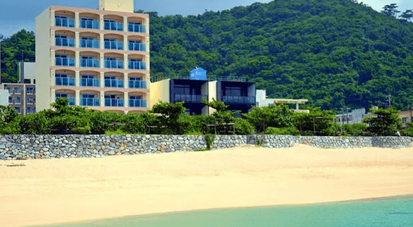 【名護住宿】Hotel Sakurano Familia Nago 外觀，建築面向海灘與山景，米白色多層樓設計，鄰近沙灘，交通便利。