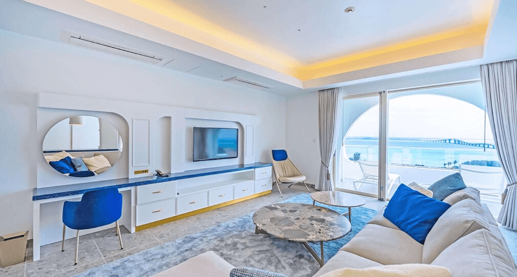 【宮古島住宿】Santorini酒店＆別墅的海景套房內部，一覽潔白牆面與藍色椅子的地中海風格客廳，窗外可見壯麗的海景與橋樑。