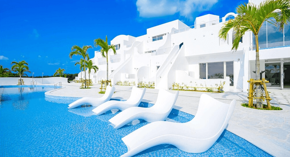 【宮古島住宿】Santorini酒店＆別墅的戶外泳池區，設有白色躺椅與希臘風白色建築，展現地中海渡假氛圍與熱帶棕櫚景致。