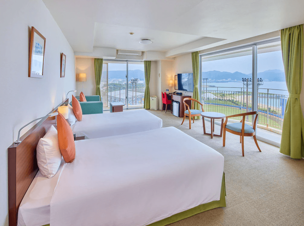 【名護住宿】"名護住宿精選示意圖"Yugaf Inn飯店沖繩 (Hotel Yugaf Inn Okinawa) 海景客房，兩張雙人床搭配柔和色系抱枕與沙發，大片落地窗外可眺望海灣與山景，房內設有電視、書桌與休憩座椅區。