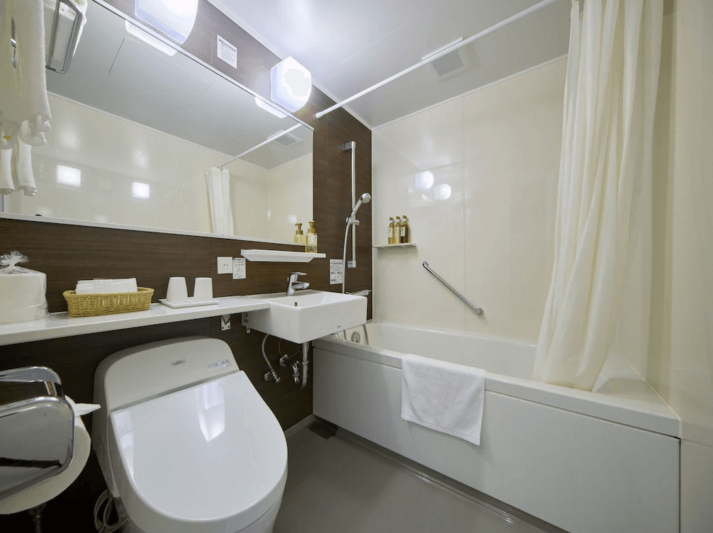 Yugaf Inn飯店沖繩 (Hotel Yugaf Inn Okinawa) 衛浴空間，配有免治馬桶、洗手台與大鏡面，浴缸旁附有洗浴用品與安全扶手，設計乾淨明亮。