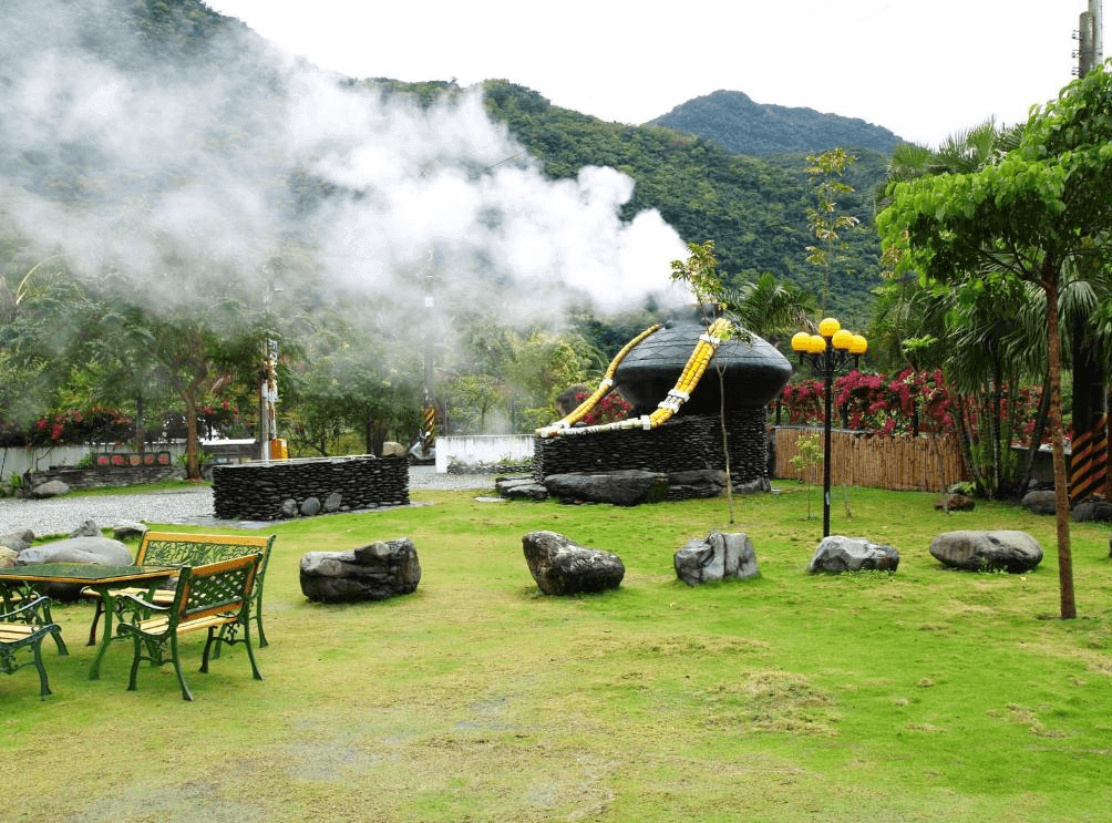 【金崙住宿】"太麻里住宿"丹堤溫泉會館戶外庭園，綠草地與石椅桌，遠處可見蒸氣騰騰的溫泉設備，背靠群山景致宜人。