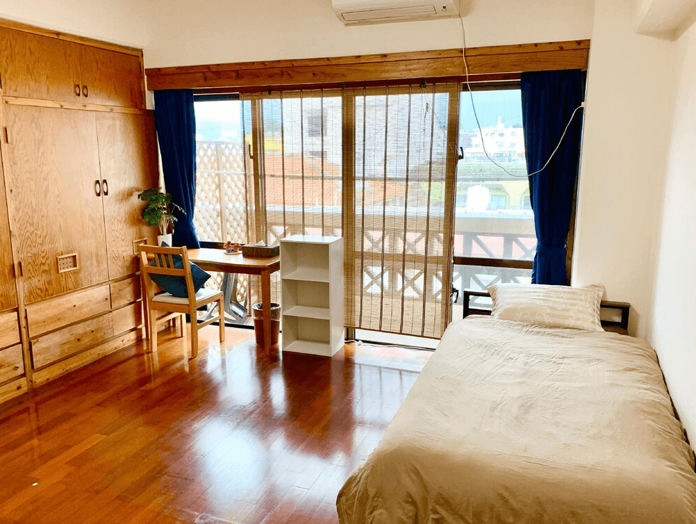 【名護住宿】全友民宿 (All Friends Guest House) 單人房，木質地板搭配單人床、書桌與椅子，落地窗外可見陽台景色，房間簡約明亮。