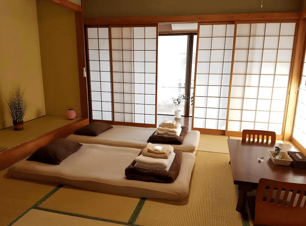 【名護住宿】全友民宿 (All Friends Guest House) 日式榻榻米房，兩組鋪設好的日式床墊，旁邊擺有茶几與木椅，拉門後方透進自然光，氛圍溫馨。