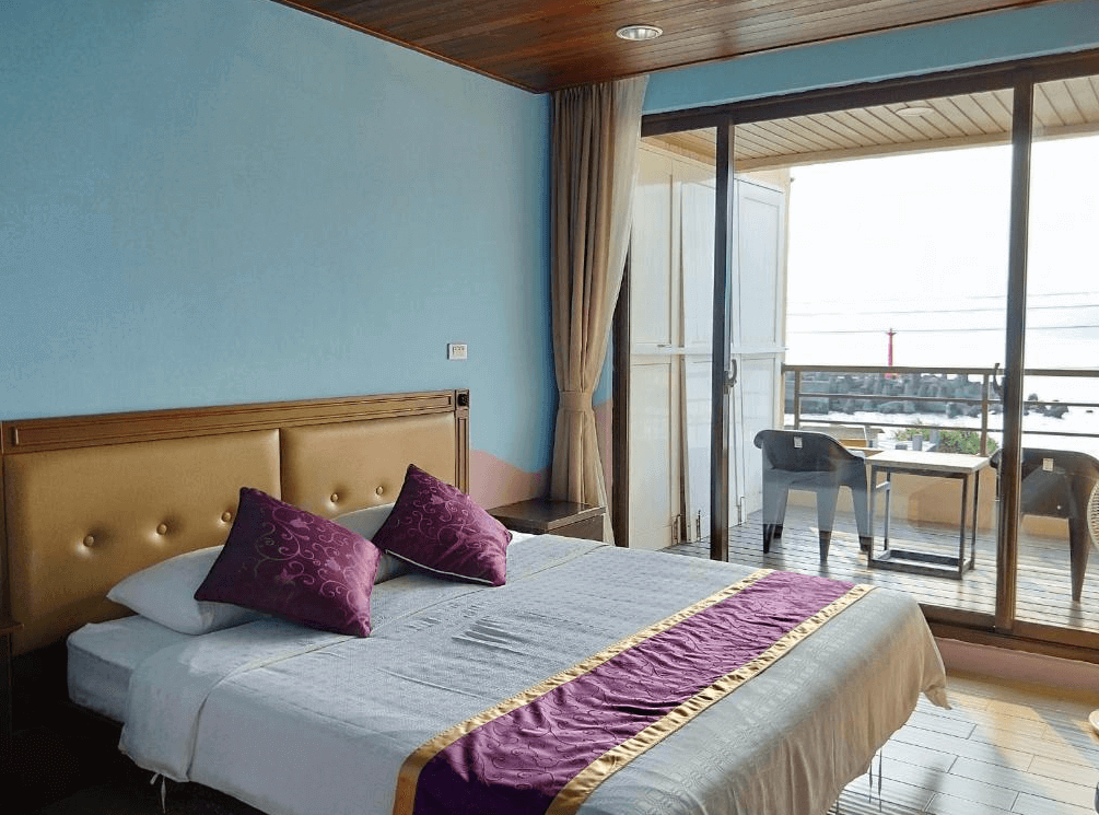 【蘭嶼住宿】兩隻魚民宿海景客房：房間設有大床與紫色抱枕，落地窗外可見陽台及海景，並配置桌椅可觀賞港灣景色。