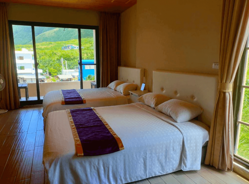 【蘭嶼住宿】兩隻魚民宿山景客房：房內擺放兩張雙人床，床尾鋪有紫色緞帶裝飾布，大片落地窗外能看到翠綠山景與村落。