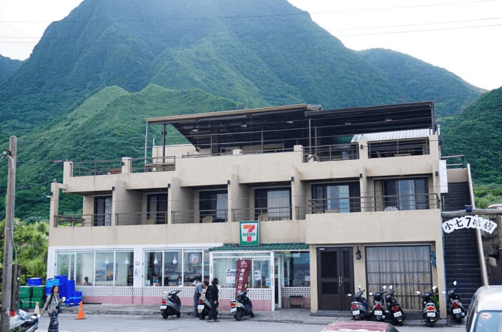 【蘭嶼住宿】兩隻魚民宿外觀：建築位於青山環抱的地段，設有多個陽台與露台，一樓附設 7-Eleven 便利商店，門口可停放機車。