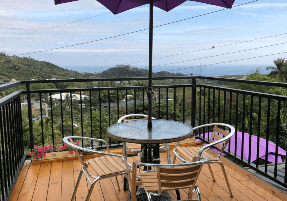 【太麻里住宿】吉廬夫敢藝文民宿觀景陽台，木質地板搭配玻璃圓桌與戶外椅，紫色遮陽傘下可遠眺山景與海景。