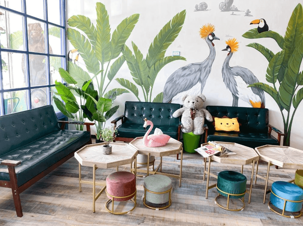 【墾丁寵物友善住宿】墾丁上好旅店大廳休憩區 — 牆面繪有熱帶植物與大型鳥類彩繪，搭配綠色皮沙發、絨布圓凳與木質六角形茶几，擺放可愛玩偶與雜誌，營造溫馨活潑氛圍。
