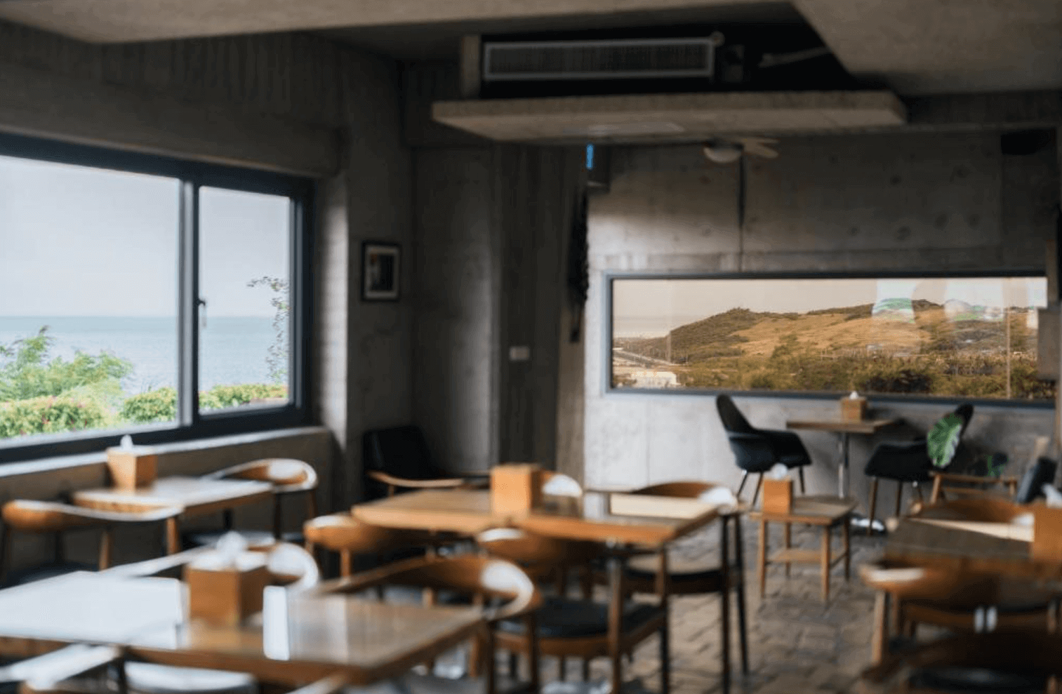 【恆春住宿】墾丁光現旅宿餐廳空間設計，大片窗景面對海岸與山丘，木質桌椅整齊擺放，沉穩灰牆與現代工業風吊燈營造出放鬆用餐環境。