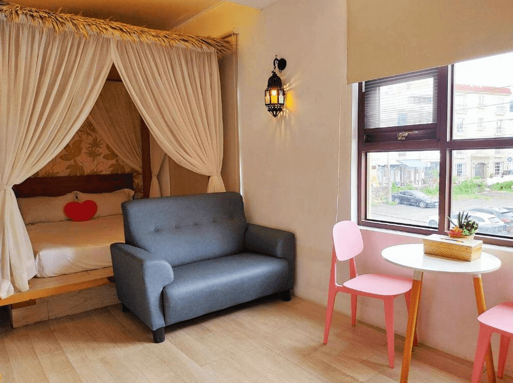 【墾丁寵物友善住宿】墾丁巴里巴里主題房 — 房間以浪漫南洋風格設計，雙人床配有白色床幔與紅色愛心抱枕，設有灰色沙發及粉色椅子與小圓桌，窗外可見街景。