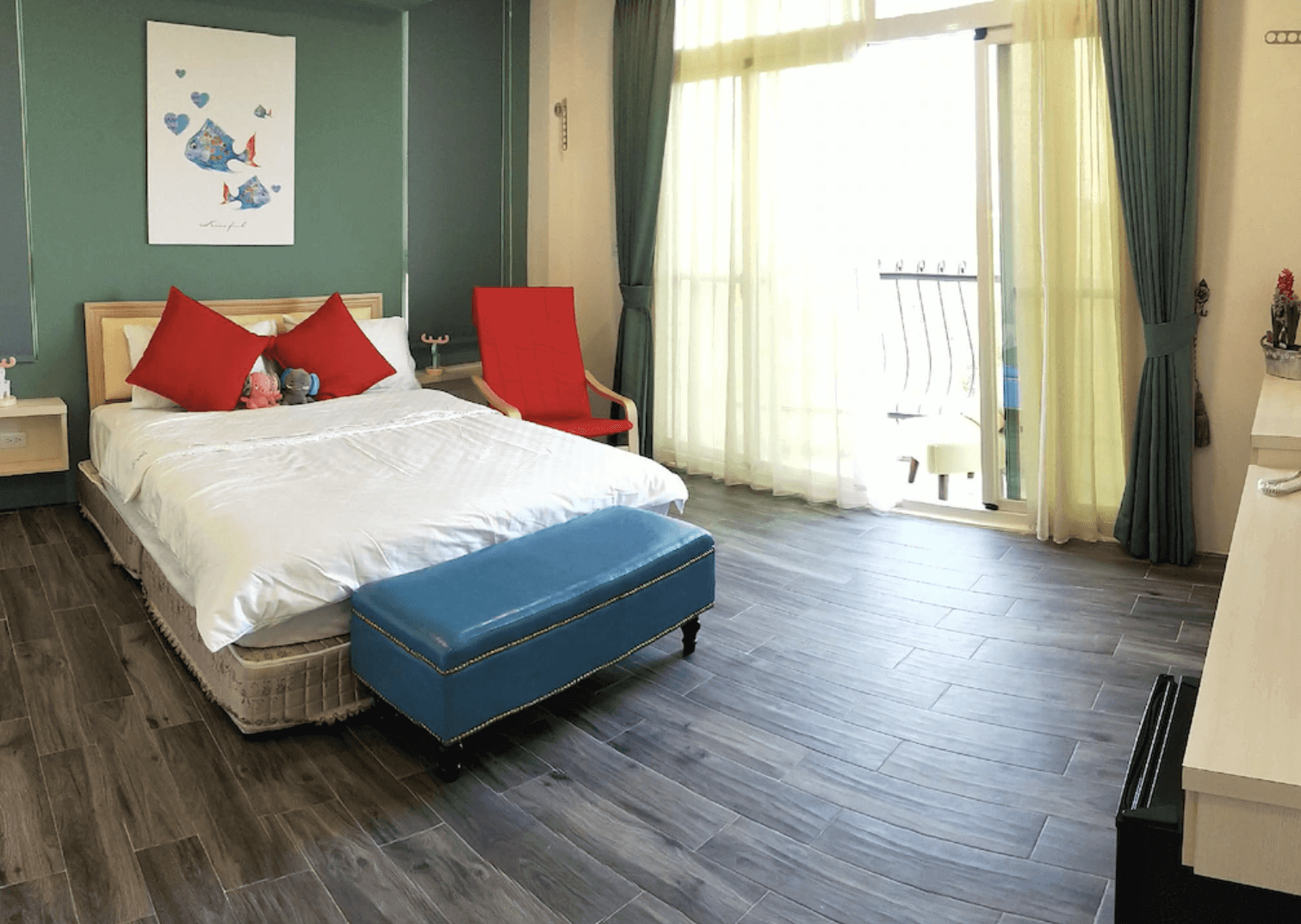 【恆春住宿】墾丁白沙灣玹玹映月民宿的雙人房型，擁有木地板與溫潤色系設計，大面窗簾後方為陽台，可遠眺戶外自然景觀，床鋪擺設整齊，紅色靠枕與椅子增添活潑感。