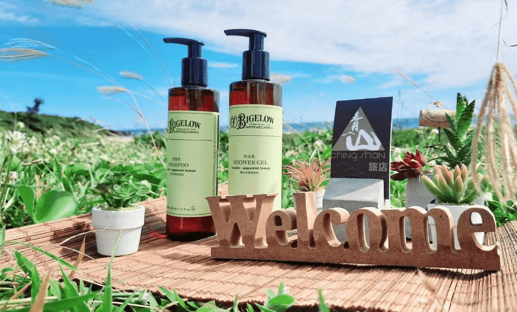 【墾丁寵物友善住宿】墾丁青山旅店迎賓擺設 — 戶外草地上擺放沐浴用品與洗髮精，搭配多肉植物與木製「Welcome」字樣裝飾，背景是藍天綠地。