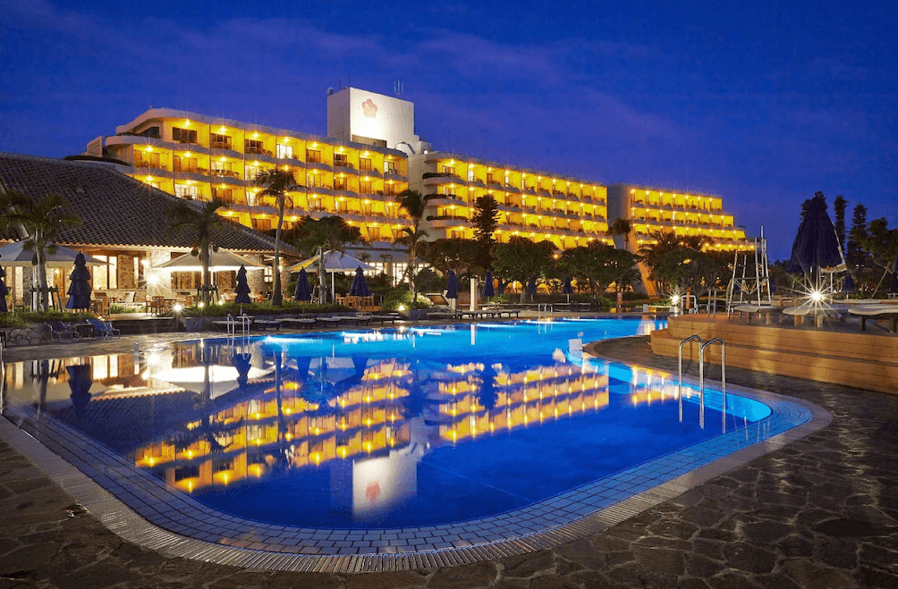 【宮古島住宿】夜晚的宮古島東急飯店全景，金黃色燈光照映在湛藍泳池中，展現迷人的熱帶度假氛圍，建築周圍環繞椰子樹與露天休息區，是享受悠閒時光的理想選擇。