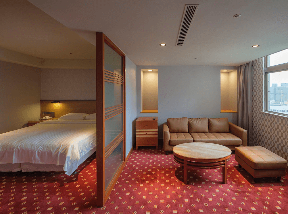 【板橋住宿】富逸旅趣-板橋府中館 精緻套房 – 寬敞的套房空間，配有雙人大床與獨立客廳區，沙發與圓桌營造休閒氛圍，大片窗戶可眺望城市景觀。