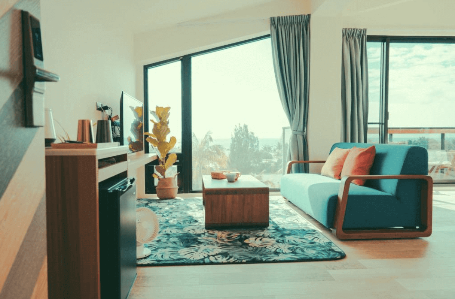 【恆春住宿】《墾丁白砂15 Villa民宿》客廳區域，配置有藍色沙發、原木茶几與大片景觀窗戶，地毯上帶有熱帶植物圖騰，整體空間明亮舒適。