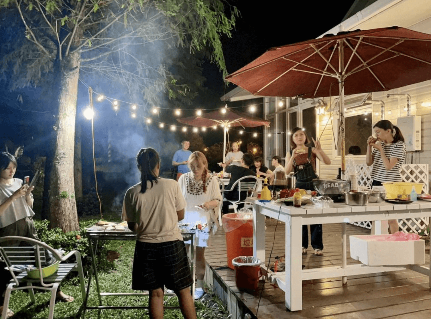【吉安住宿】干城小鎮民宿 戶外BBQ活動 — 夜間庭院烤肉聚會，燈串與紅色陽傘營造熱鬧氛圍，旅客們一同享用美食、互動交流。