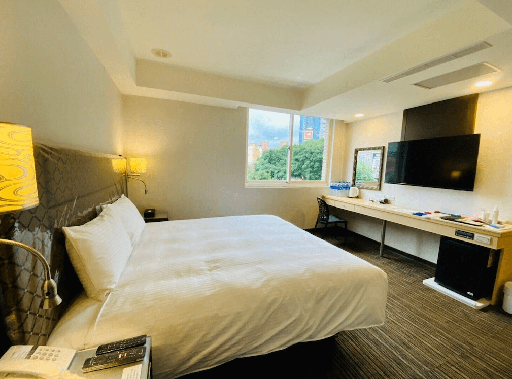 【板橋住宿】旅居文旅板橋車站館客房一：現代簡約設計的大床房，設有大片窗戶可眺望綠意與市景，房內配備電視、書桌、熱水壺與瓶裝水，空間明亮舒適。