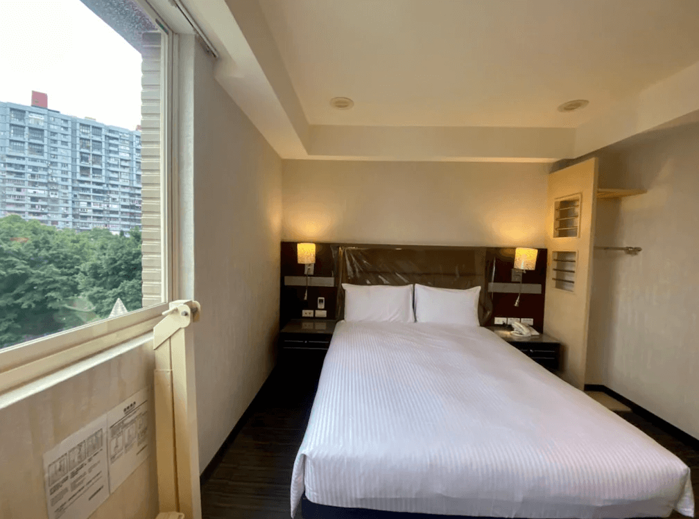 【板橋住宿】旅居文旅板橋車站館客房二：溫馨舒適的大床房，床頭兩側附有閱讀燈與床邊桌，窗外可見綠樹與城市建築，整體氛圍簡單乾淨。