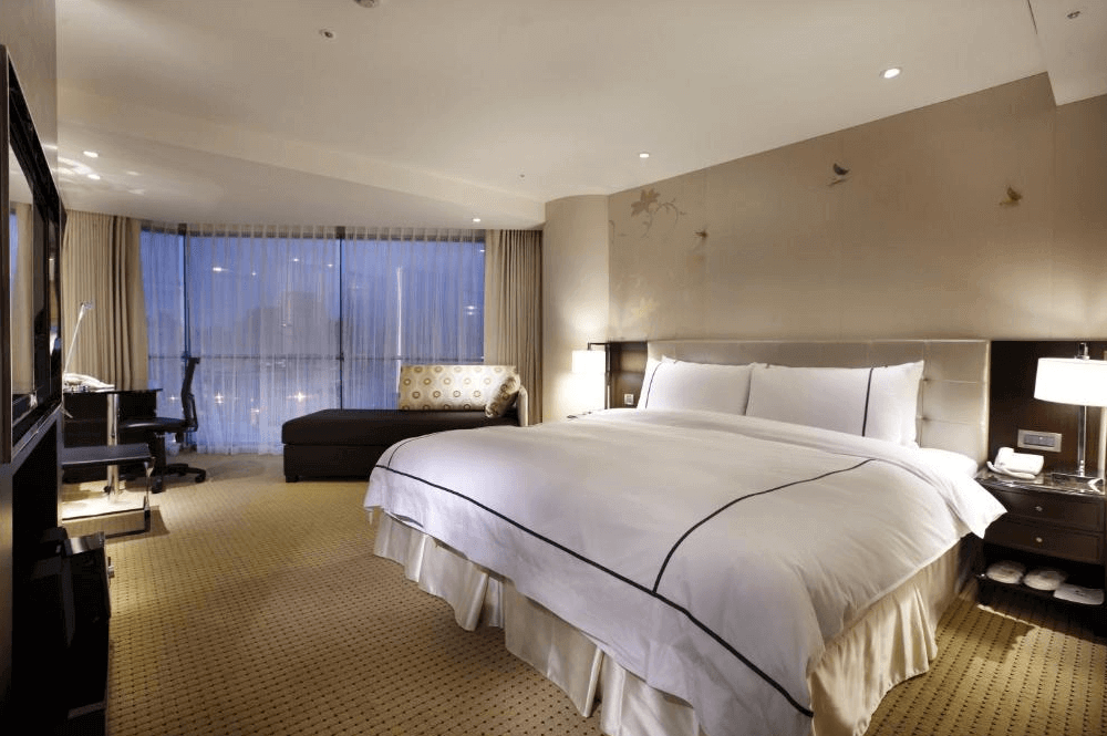 【大安住宿】柯達大飯店台北敦南豪華客房 —— 寬敞舒適的雙人房，大床搭配白色寢具，房內設有沙發、書桌與大片落地窗，可欣賞城市夜景。