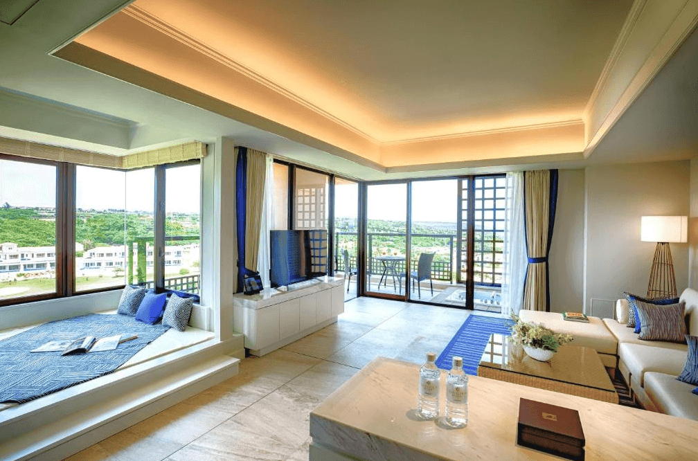 【宮古島住宿】汐姬海市飯店的豪華套房設有窗邊臥榻閱讀區與寬敞客廳，三面環窗引入自然光與綠意景觀，展現高雅放鬆的住宿體驗。