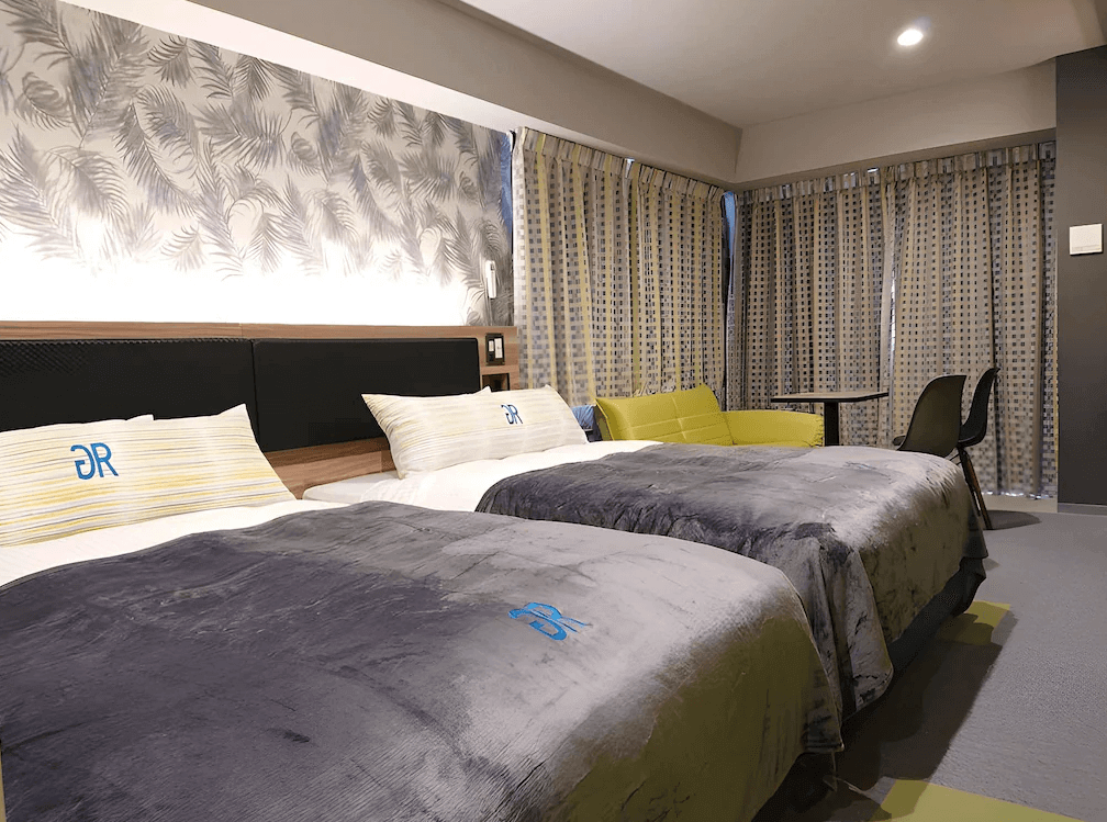 【名護住宿】沖繩名護人工溫泉二股湯之華格林里奇飯店 雙床客房，柔和燈光搭配灰藍色羽毛壁紙設計，設有兩張舒適雙人床、沙發與圓桌，空間寬敞現代。