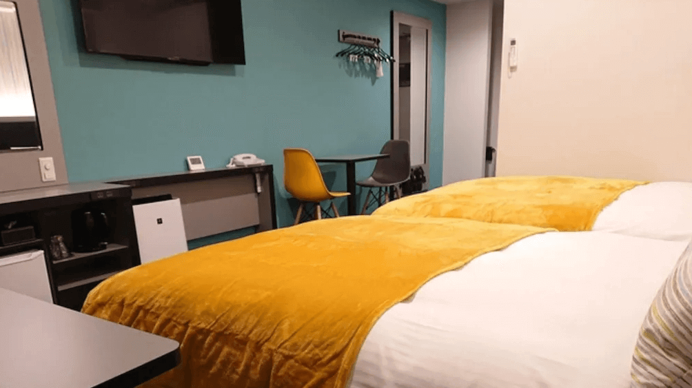 【名護住宿】沖繩名護人工溫泉二股湯之華格林里奇飯店 雙人客房，床上配有亮黃色毛毯，房內設有電視、冰箱、書桌與多功能座椅，裝潢簡約明亮。
