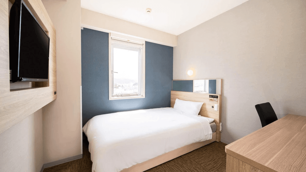【名護住宿】沖繩名護超級酒店 (Super Hotel OKINAWA NAGO) 單人房，設有簡約單人床、木質書桌與牆掛式電視，窗外可見市景，空間整潔明亮。