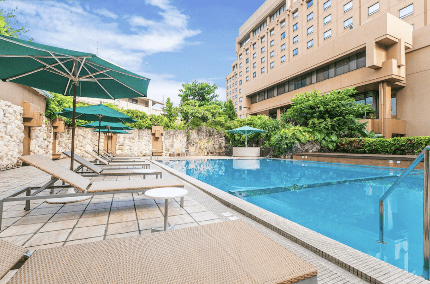 【那霸住宿】沖繩海景飯店室外泳池 — 飯店外的藍色泳池旁設有綠色遮陽傘與躺椅，周圍綠植與飯店建築營造度假氛圍。