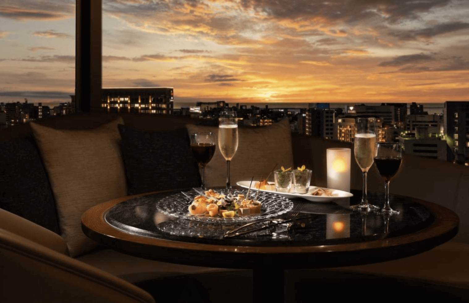 【那霸住宿】沖繩海景飯店夕陽餐桌 — 露台餐桌擺放著氣泡酒、紅酒與精緻餐點，背景是夕陽映照下的城市與遠方海平面。