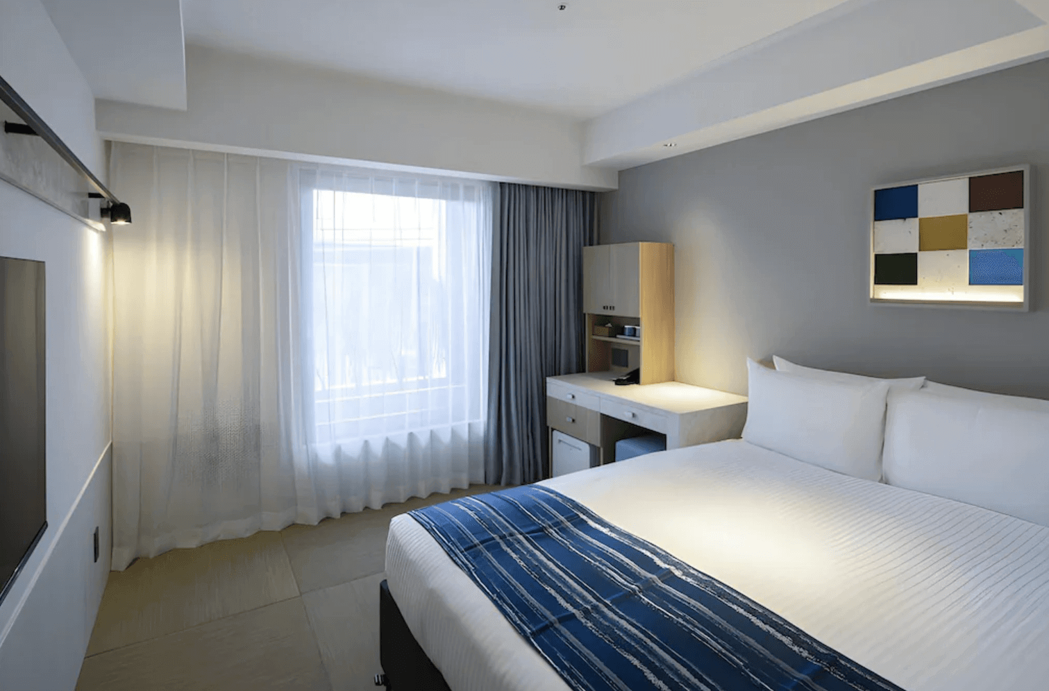 【那霸住宿】沖繩逸之彩飯店標準客房 — 一張雙人床搭配藍色床旗，簡約現代設計，內有書桌與大面窗簾引入自然光。