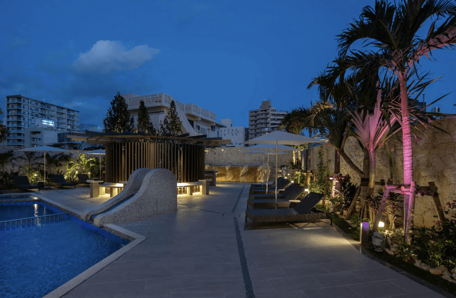 【那霸住宿】沖繩逸之彩飯店戶外泳池 — 夜間燈光映照的泳池區，設有滑梯、躺椅與遮陽傘，周圍種滿熱帶植栽。