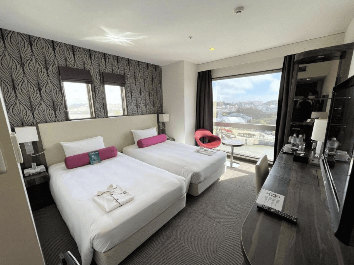 【那霸住宿】沖繩那霸美居飯店客房 — 兩張單人床搭配紫色長抱枕與淺色床頭板，牆面為深色葉紋壁紙，落地窗外可見城市與山景。
