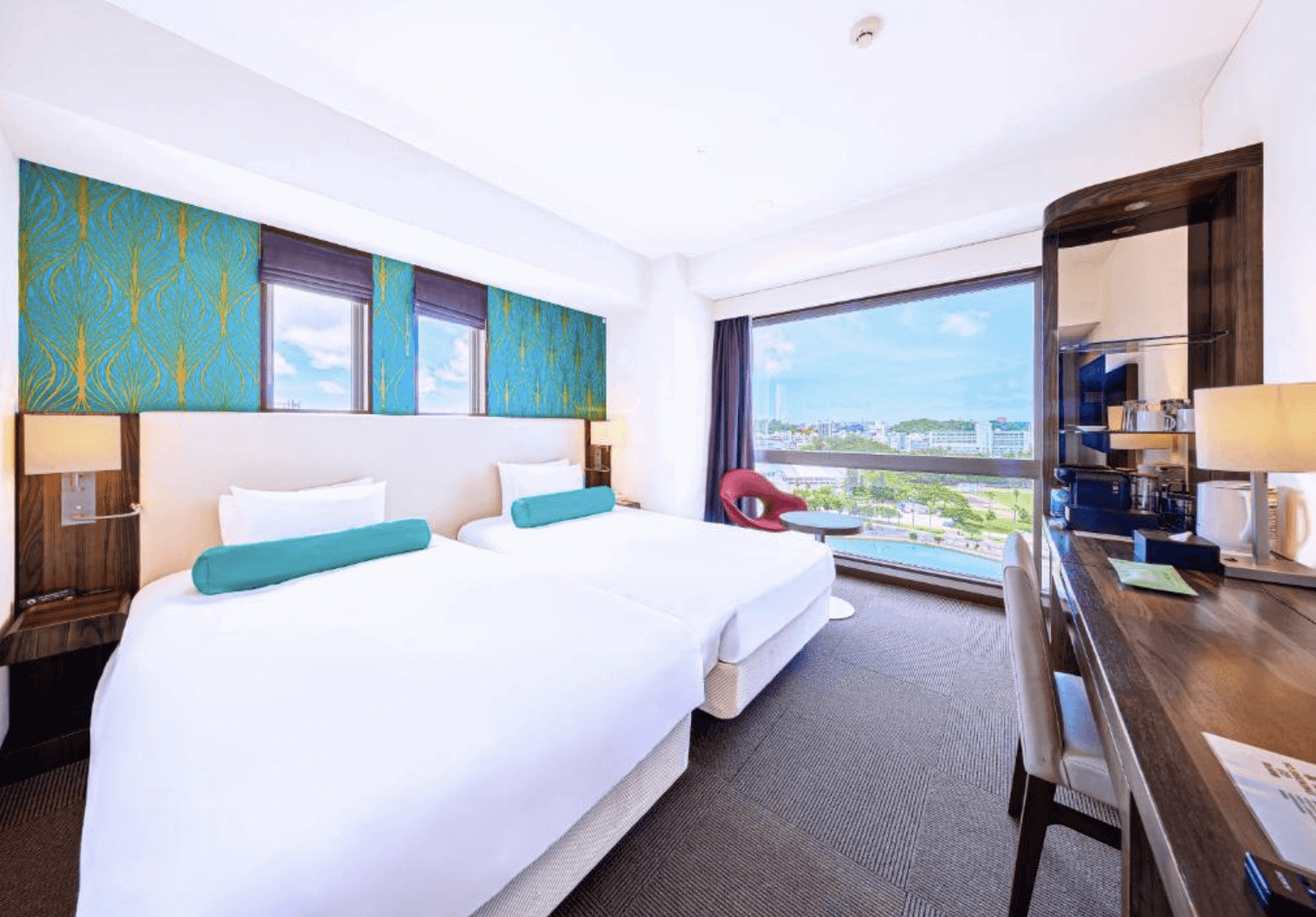 【那霸住宿】沖繩那霸美居飯店明亮客房 — 兩張單人床配藍綠色床頭牆與藍綠色長抱枕，大面落地窗外是市區與泳池景觀。
