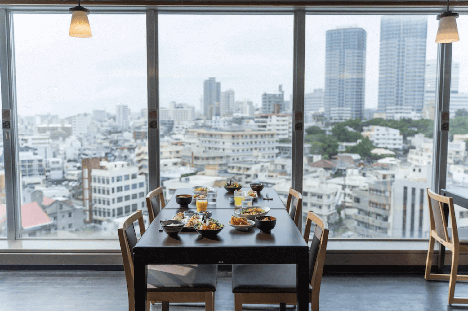 【那霸住宿】海洋飯店高樓景觀餐廳 — 餐桌擺滿日式餐點與飲品，落地窗外可俯瞰那霸市區與高樓景色。