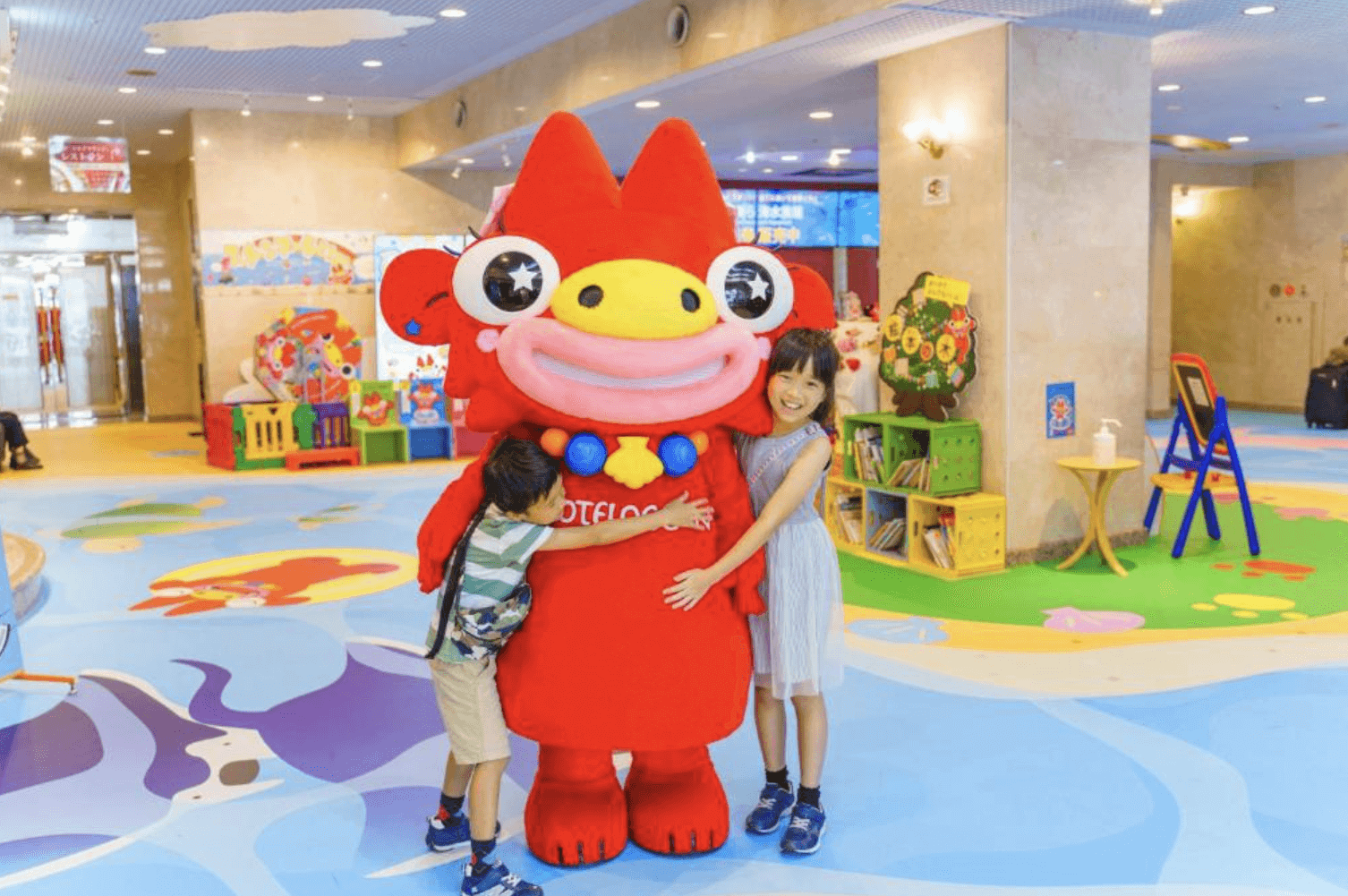 【那霸住宿】海洋飯店大廳親子互動區 — 孩子們擁抱紅色吉祥物公仔，背景為色彩繽紛的遊戲與閱讀空間。
