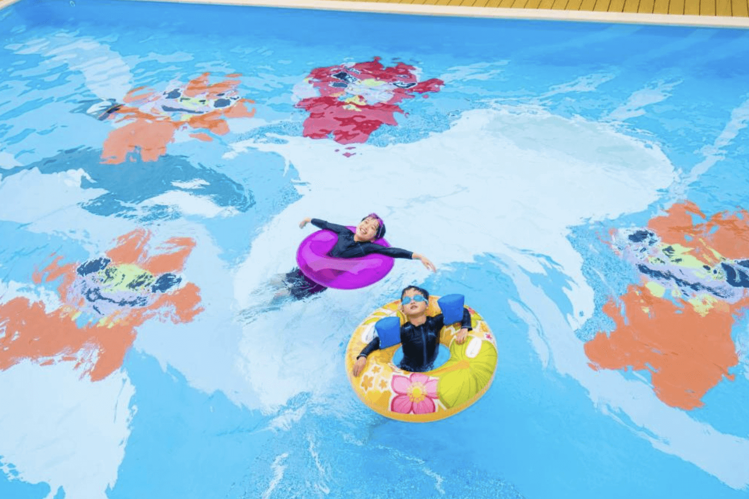 【那霸住宿】海洋飯店戶外泳池 — 兩名兒童在泳池中使用彩色游泳圈漂浮，池底印有卡通圖案，氣氛歡樂。