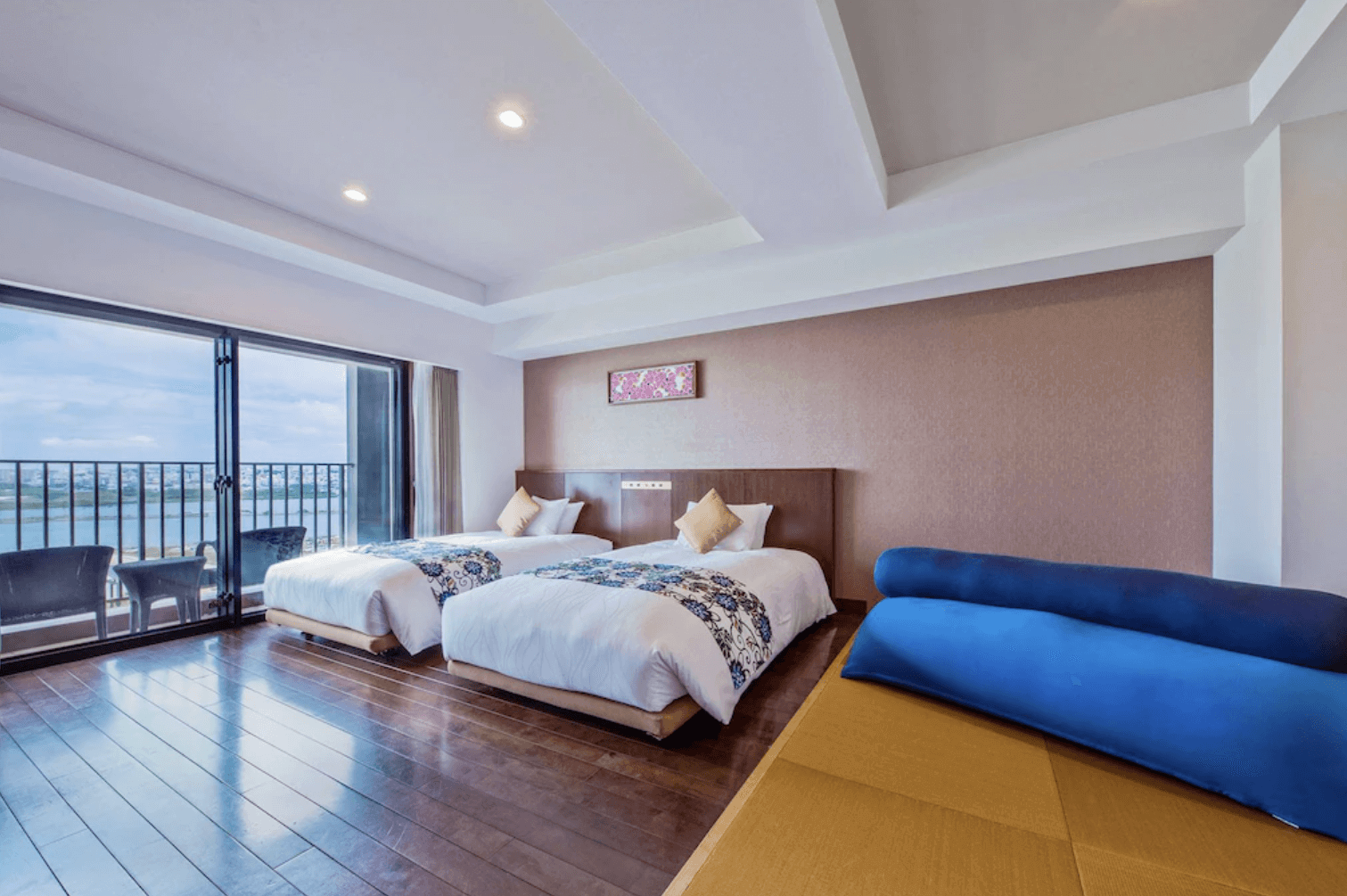 【那霸住宿】琉球溫泉瀨長島飯店客房 — 兩張單人床搭配花紋床旗與米色抱枕，旁邊設有藍色長抱枕的榻榻米區，落地窗外可見陽台與海景。