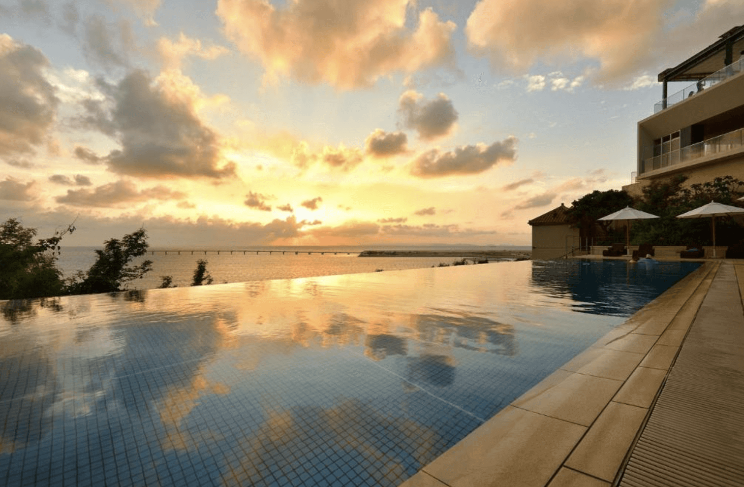 【那霸住宿】琉球溫泉瀨長島飯店無邊際泳池 — 夕陽映照下的泳池與大海相連，遠方可見跨海大橋與金黃色天空，充滿浪漫氛圍。