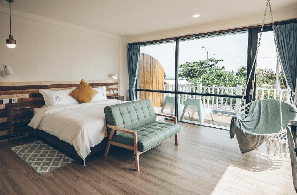 【墾丁寵物友善住宿】草分木民宿海景房 — 木質地板搭配溫暖色調設計，配置一張雙人床、綠色沙發與吊掛式鞦韆椅，大面落地窗可通往陽台並欣賞戶外綠意景觀。
