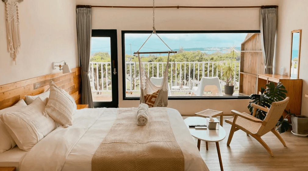 【墾丁寵物友善住宿】草分木民宿陽台海景房 — 明亮溫馨的雙人房，採用木質家具與米色系軟裝，窗外與陽台可遠眺海景，房內設有吊掛式吊椅與小茶几。