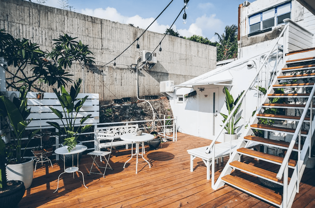 【墾丁寵物友善住宿】草分木民宿戶外露台 — 木質地板的開放式露台區，配有白色桌椅與綠植盆栽，樓梯可通往上層空間，環境陽光明亮且充滿度假氛圍。