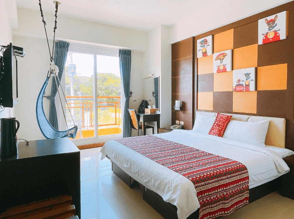 【墾丁寵物友善住宿】華納小筑雙人房 — 明亮房型內設有一張雙人床，床頭以橘棕色方格拼接設計，牆上掛有卡通狗插畫，房內配置藍色吊椅與辦公桌，落地窗外可見陽台景色。