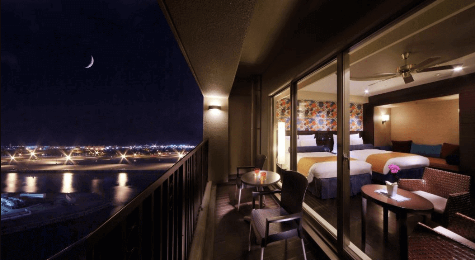 【那霸住宿】那霸遙索爾Spa中心酒店夜景陽台 — 陽台擺放桌椅，可欣賞夜晚機場燈光與彎月景色，透過玻璃可見溫馨客房內的兩張雙人床與裝飾牆。