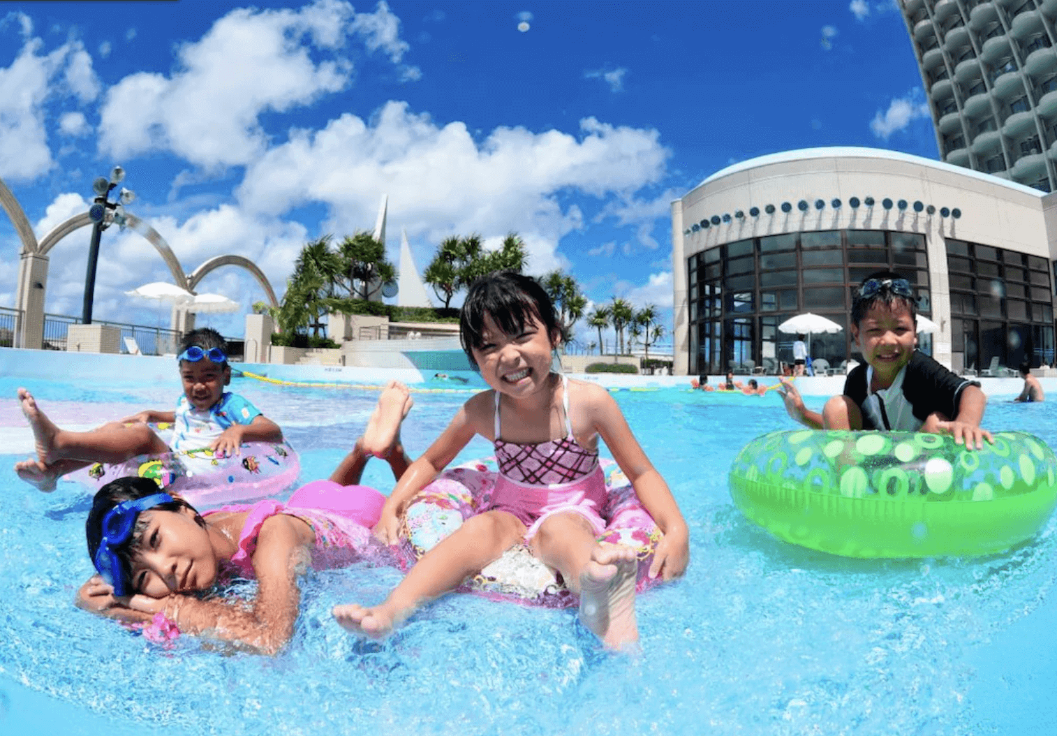 【那霸住宿】那霸遙索爾Spa中心酒店戶外泳池 — 藍天下兒童在泳池中嬉戲，背景為酒店建築與棕櫚樹，呈現熱帶度假氛圍。