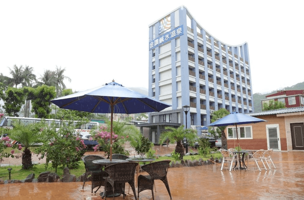 【金崙住宿】金讚溫泉飯店建築外觀，藍白相間的高樓外牆設計，前方庭院擺放戶外桌椅與遮陽傘，周圍綠意環繞。