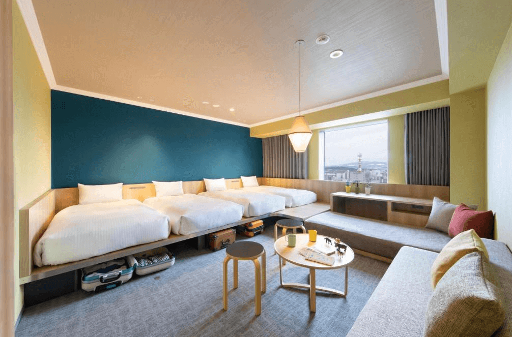 【旭川住宿】OMO7 旭川 by 星野集團多人房型，四張單人床整齊排列，大片窗戶可遠眺旭川市景，並設有沙發區與簡約木質茶几，適合家庭或團體入住。