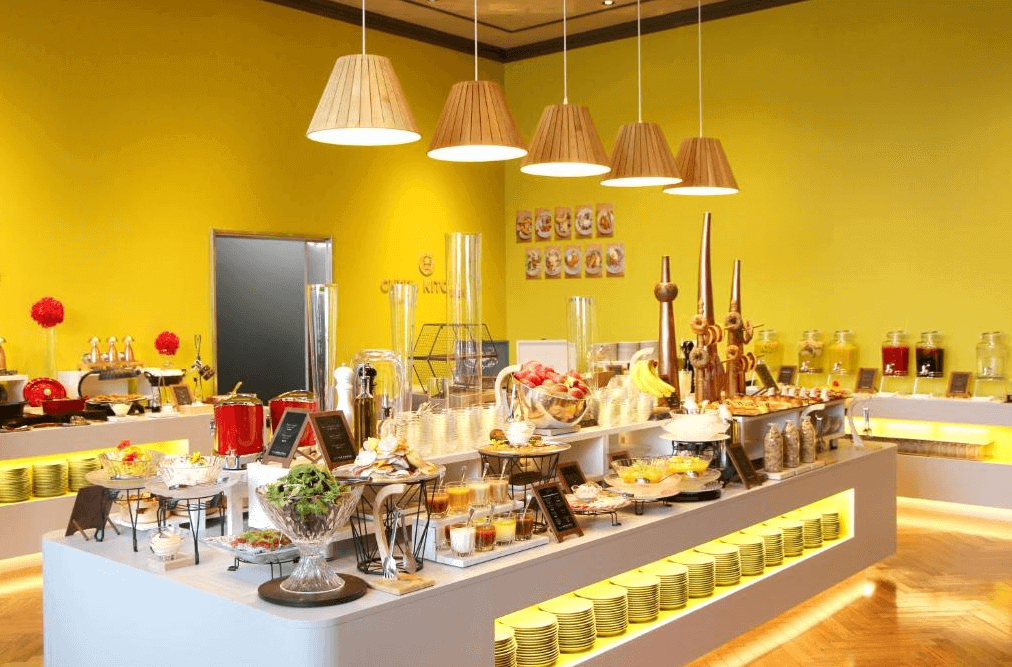 【旭川住宿】OMO7 旭川 by 星野集團早餐自助餐區，黃色牆面明亮活潑，餐檯上擺放各式水果、沙拉、甜點與飲品，提供豐盛的早餐選擇。