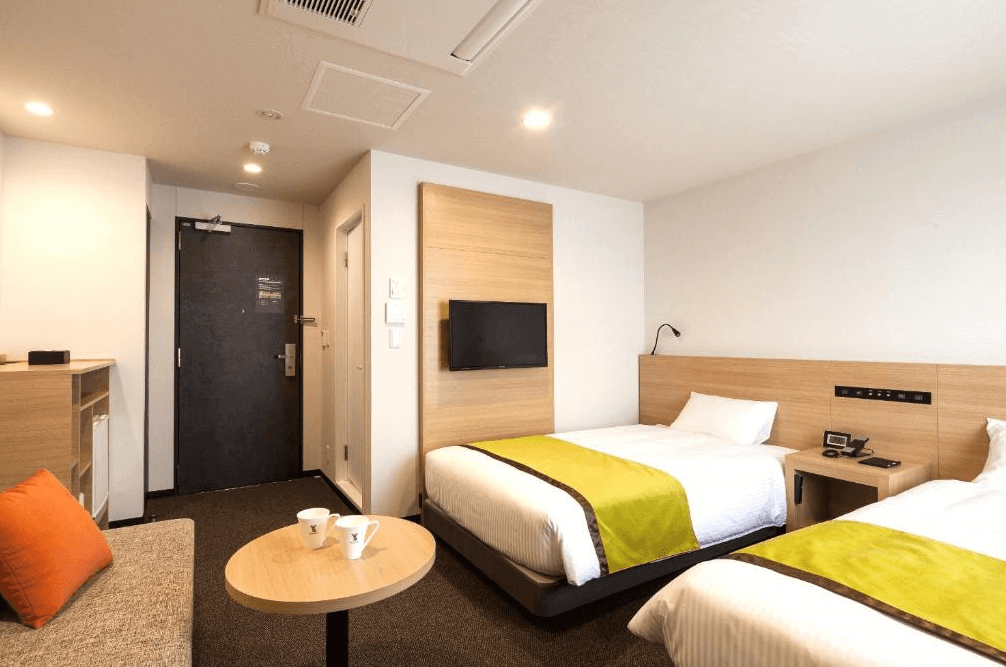 【旭川住宿】Y飯店-旭川站前 (Y's Hotel Asahikawa) 標準雙床房，簡約木質設計，兩張單人床搭配綠色床旗，房內配有電視、沙發與小茶几，氛圍溫馨舒適。